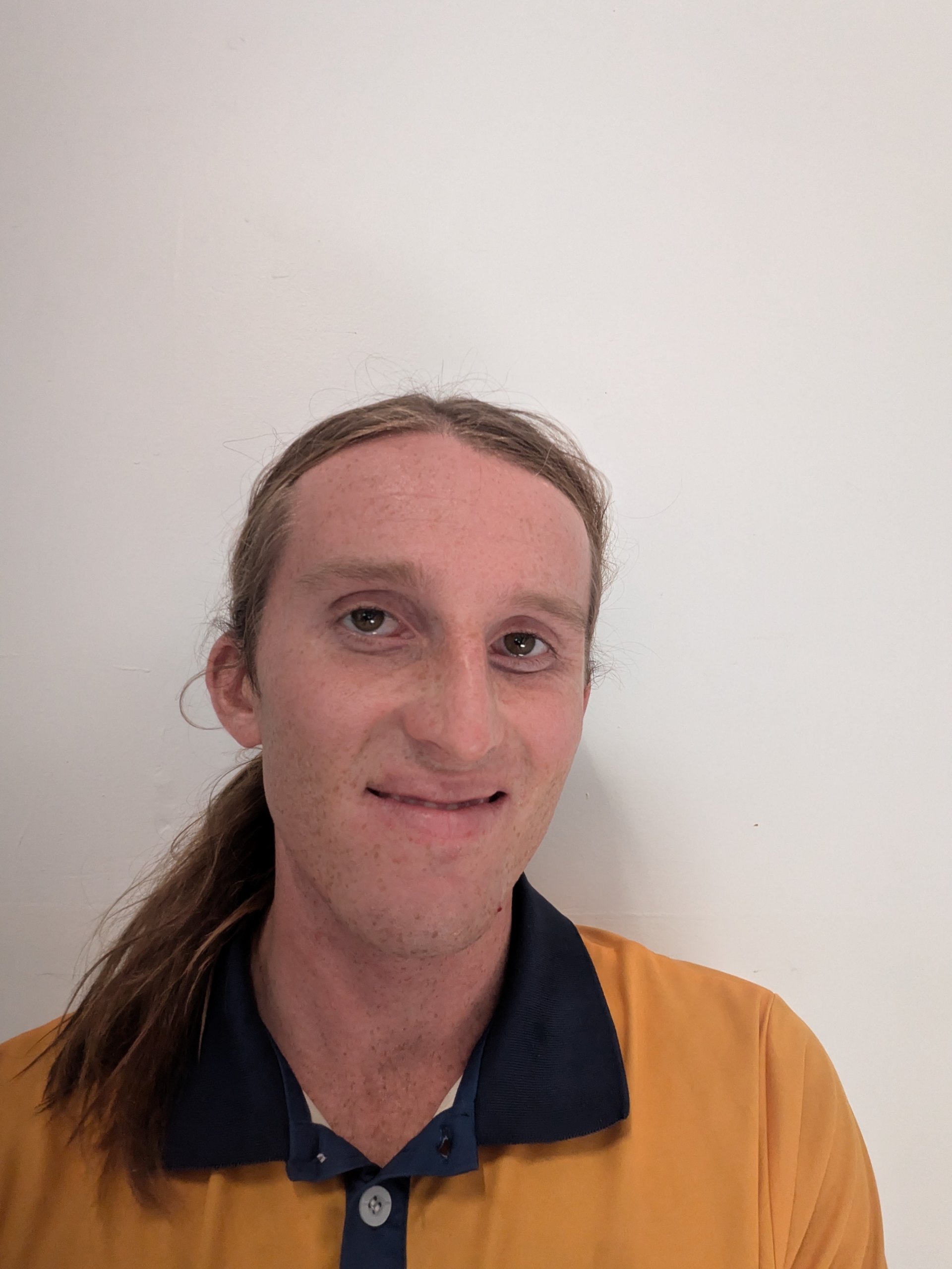 Close-up portrait of a person with long hair, wearing an orange polo with a dark navy collar, smiling at the camera against a plain light wall.