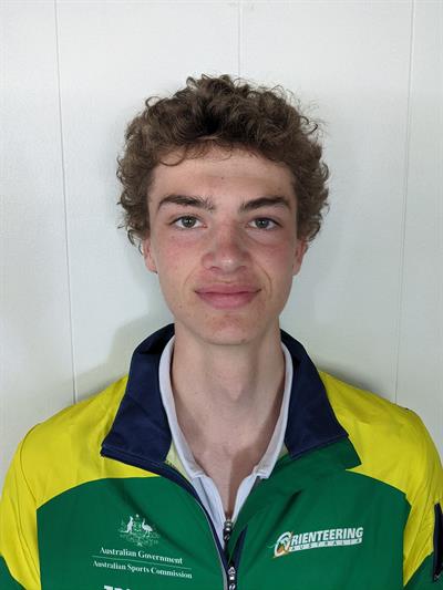 Young man with curly brown hair wearing a green and yellow sports jacket, standing against a white paneled wall.