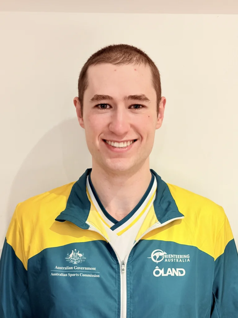 Smiling young man with short brown hair wearing a teal and yellow sports jacket with Australian Government and Orienteering Australia logos, facing the camera.