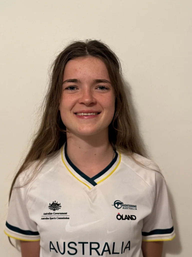 Smiling young woman with long hair in a white Australia sports jersey with teal and yellow trim and sponsor logos on the chest area, facing camera.