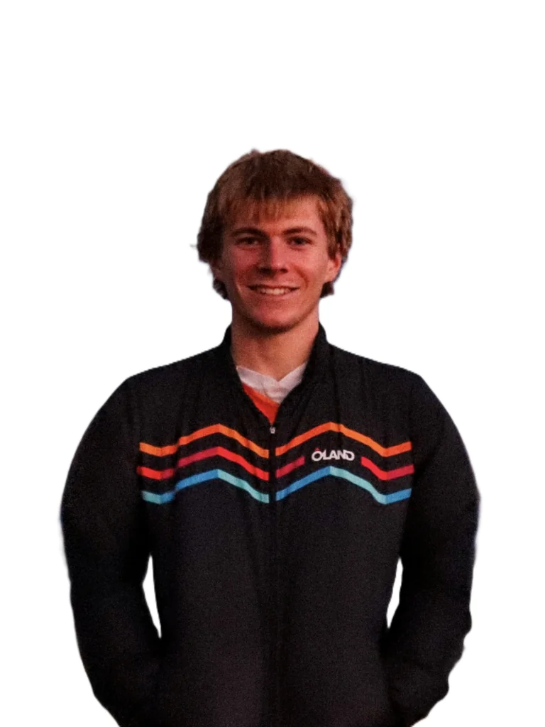 Young man with short blond hair wearing a black sweater with colorful chevron stripes and a small logo on the chest, posed against a white background.