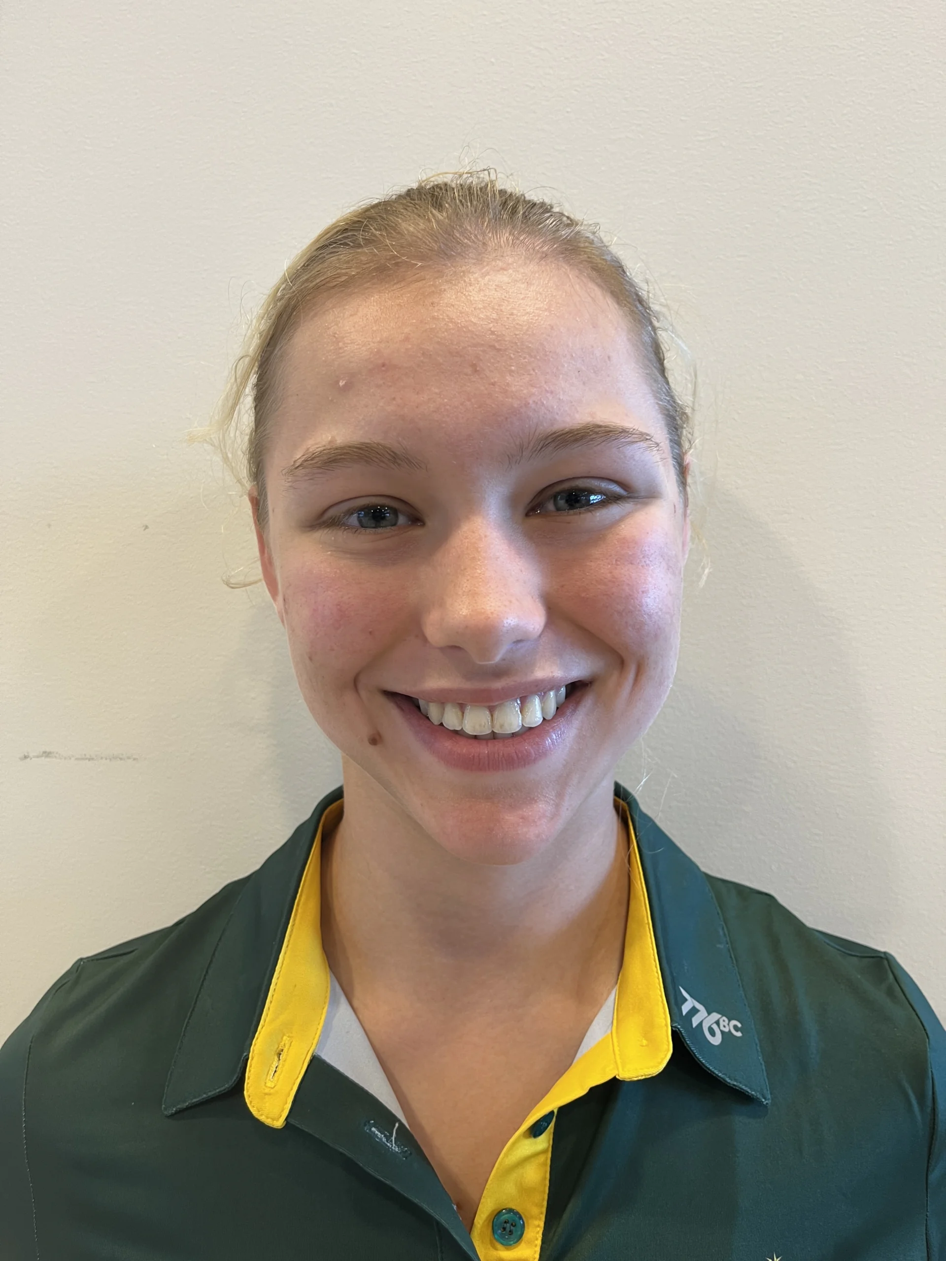 Close-up portrait of a smiling person wearing a green and yellow uniform against a plain wall.