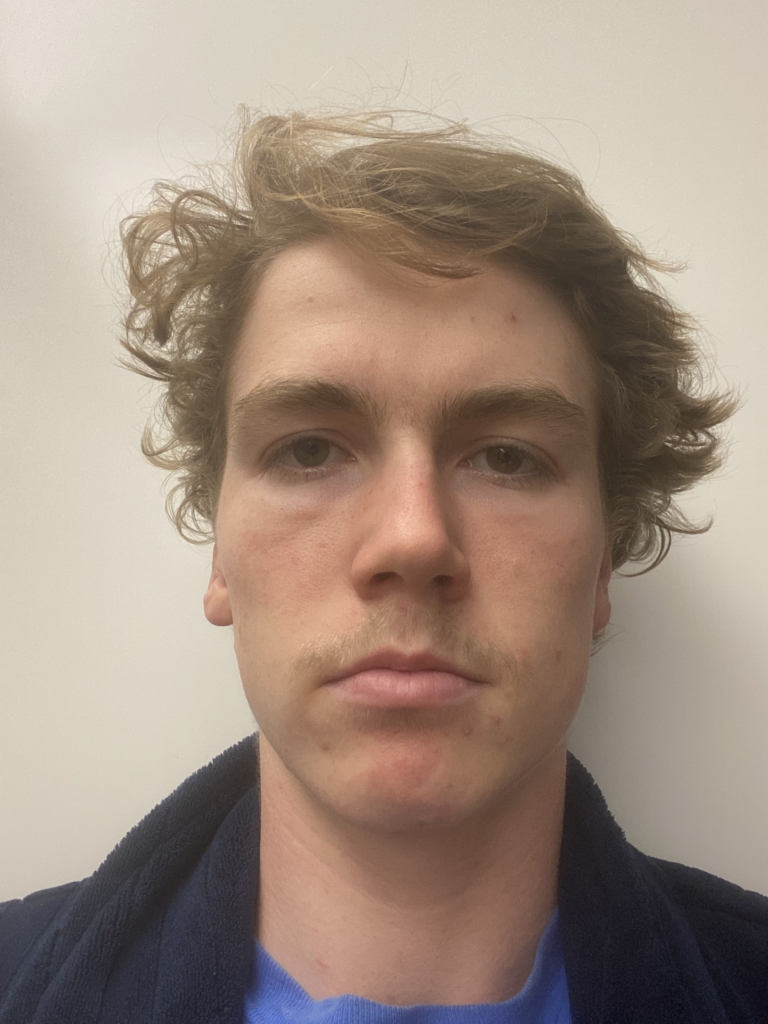 Close-up portrait of a young man with tousled blond hair, wearing a blue shirt and dark sweater.