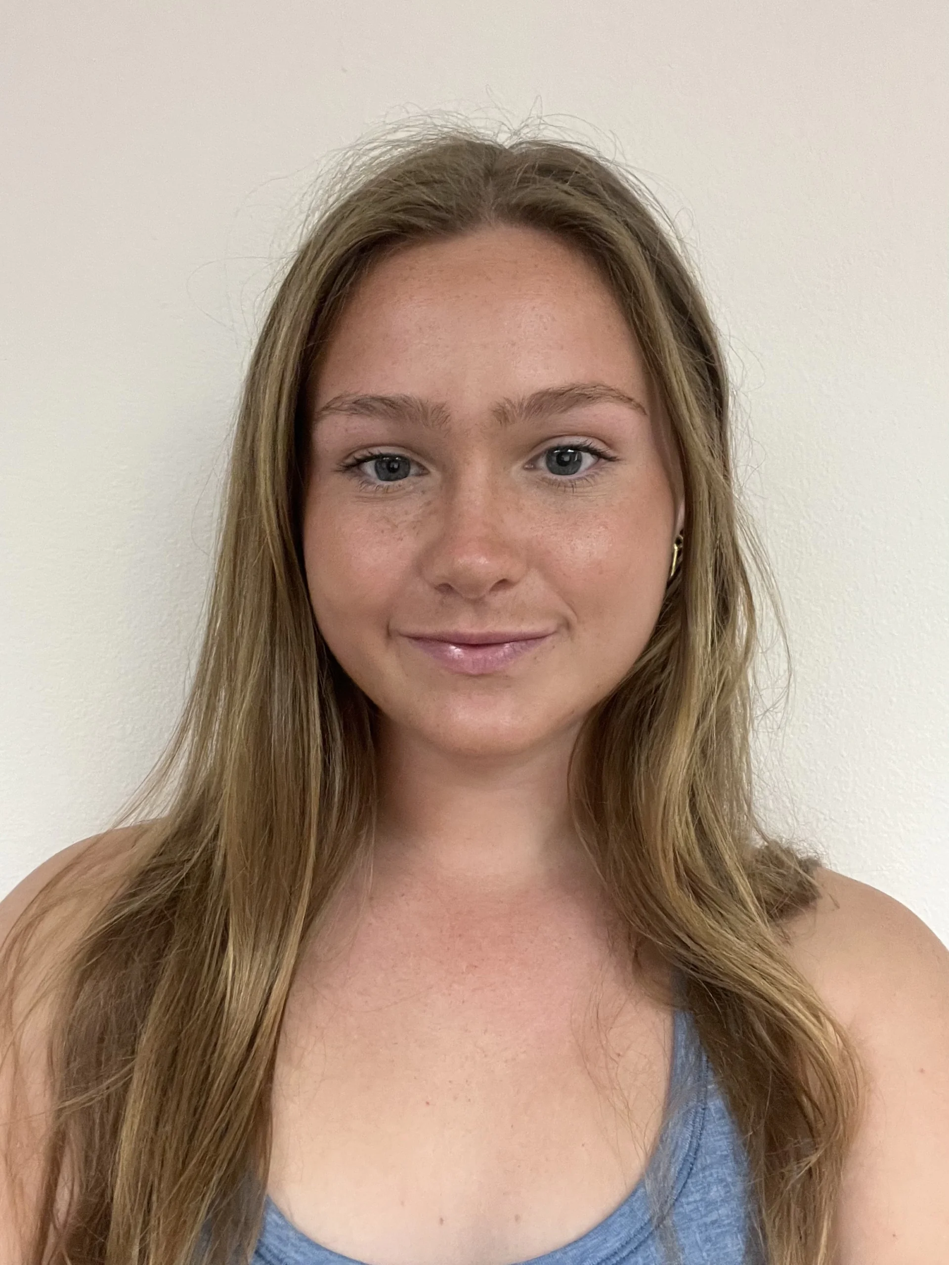 Head-and-shoulders portrait of a young woman with long light brown hair and a blue top against a beige wall.