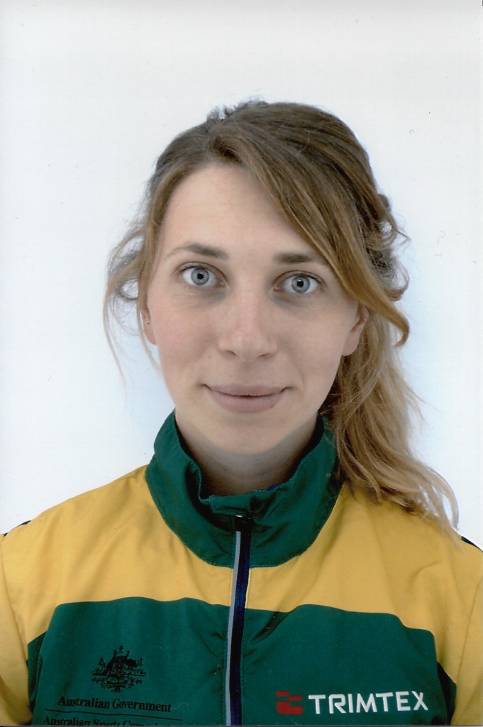 Close-up portrait of a woman with blue eyes and light brown hair, wearing a green-and-yellow Trimtex jacket.
