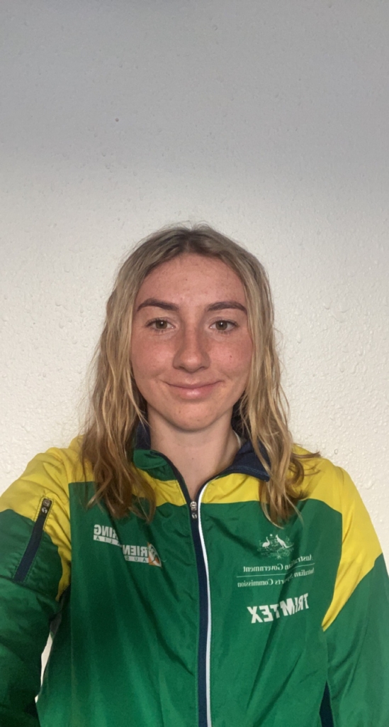 Smiling young woman with shoulder-length blond hair wearing a green and yellow jacket, standing against a white textured wall.
