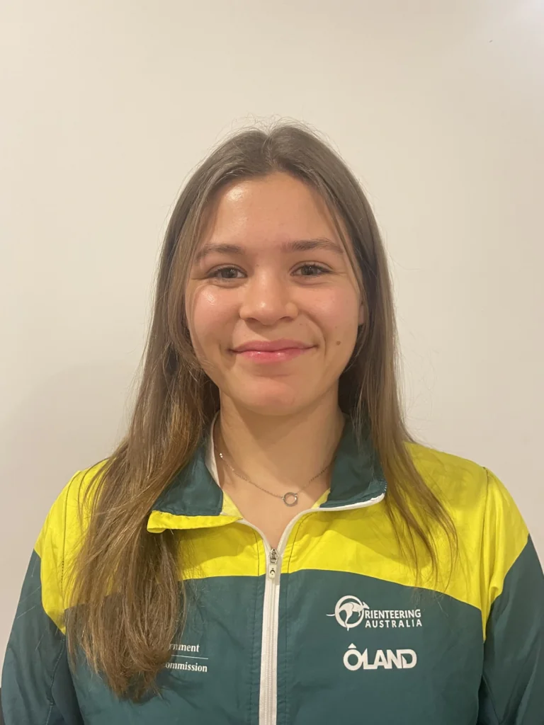 Portrait of a smiling woman with long brown hair wearing a green-and-yellow outdoor jacket with logos on the chest (Orienteering Australia).