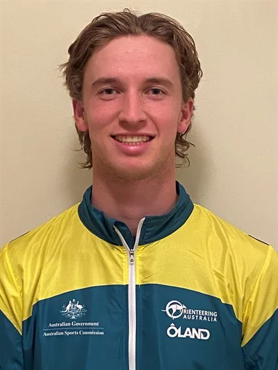 Smiling young man wearing a yellow and blue sports jacket with logos, standing against a plain beige wall.