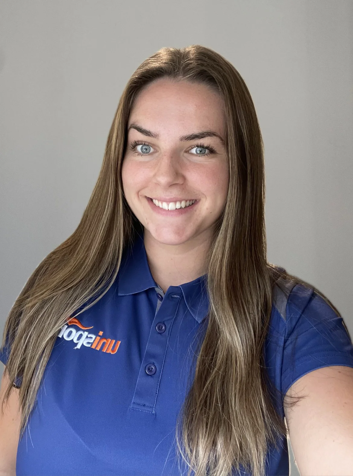 Smiling woman with long light brown hair in a blue polo shirt against a neutral gray background.
