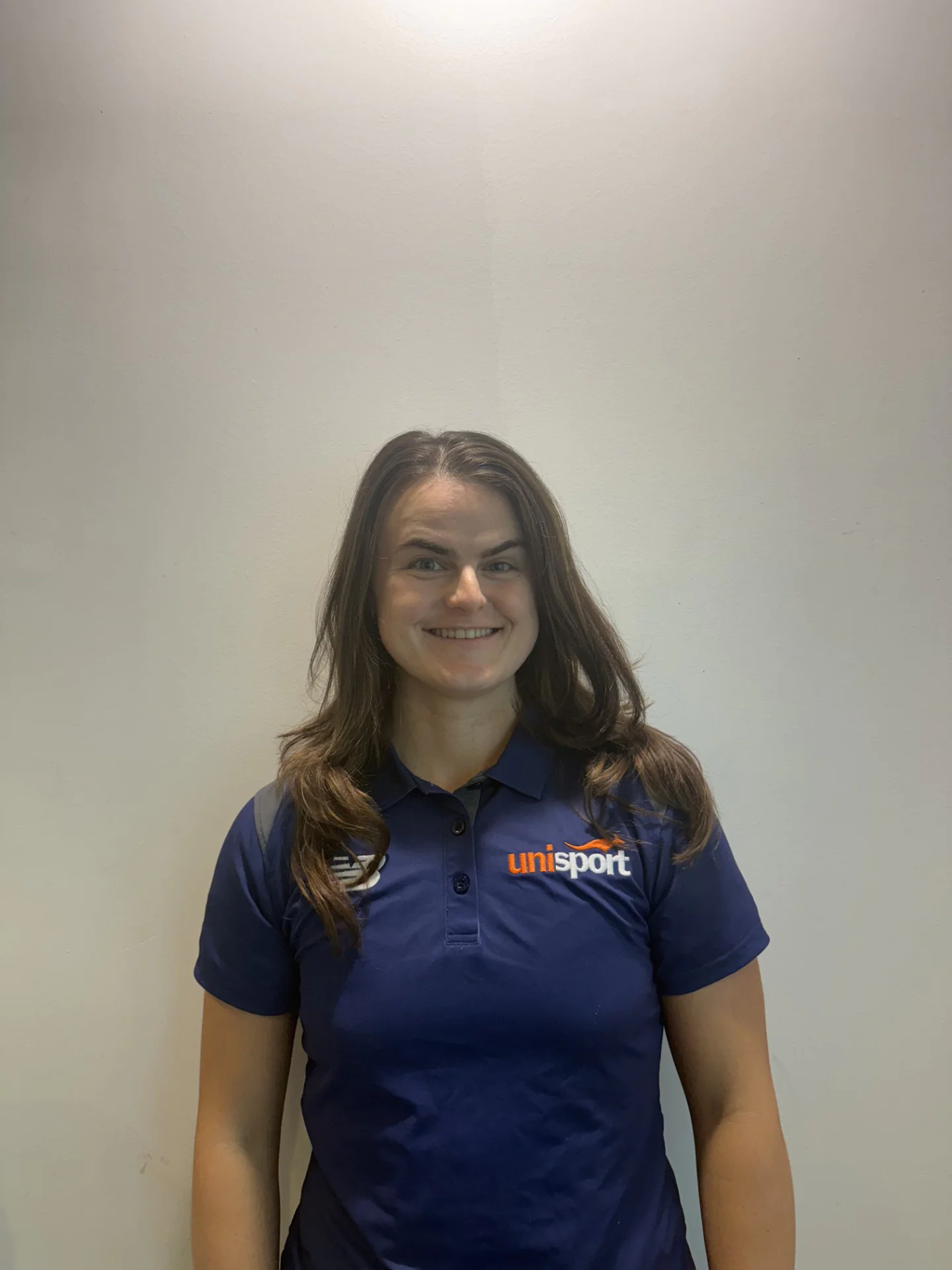 Smiling young woman with long brown hair wearing a dark blue polo shirt with a uniSport logo, standing against a plain beige wall.