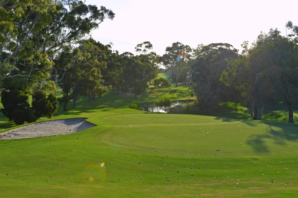 Golf course green with a sand bunker on the left, trees framing the scene and a flag near the hole by a small pond beyond.