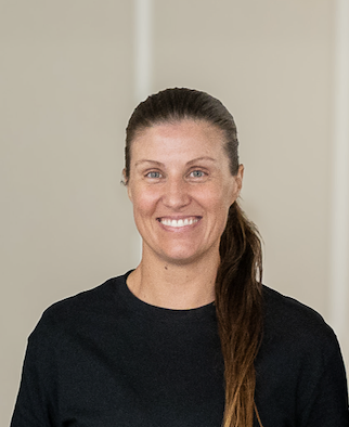 Smiling woman with long brown ponytail wearing a black shirt, standing indoors.