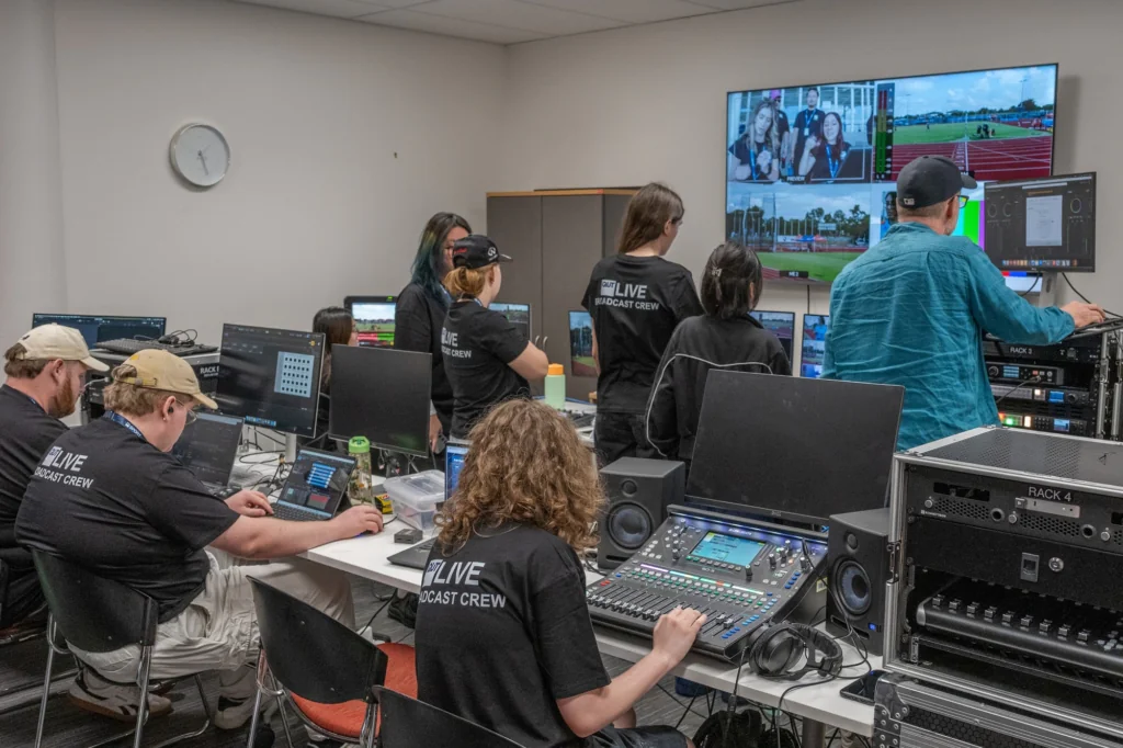 Broadcast control room with crew at desks using laptops and mixing consoles; large wall monitor shows multiple sports video feeds.
