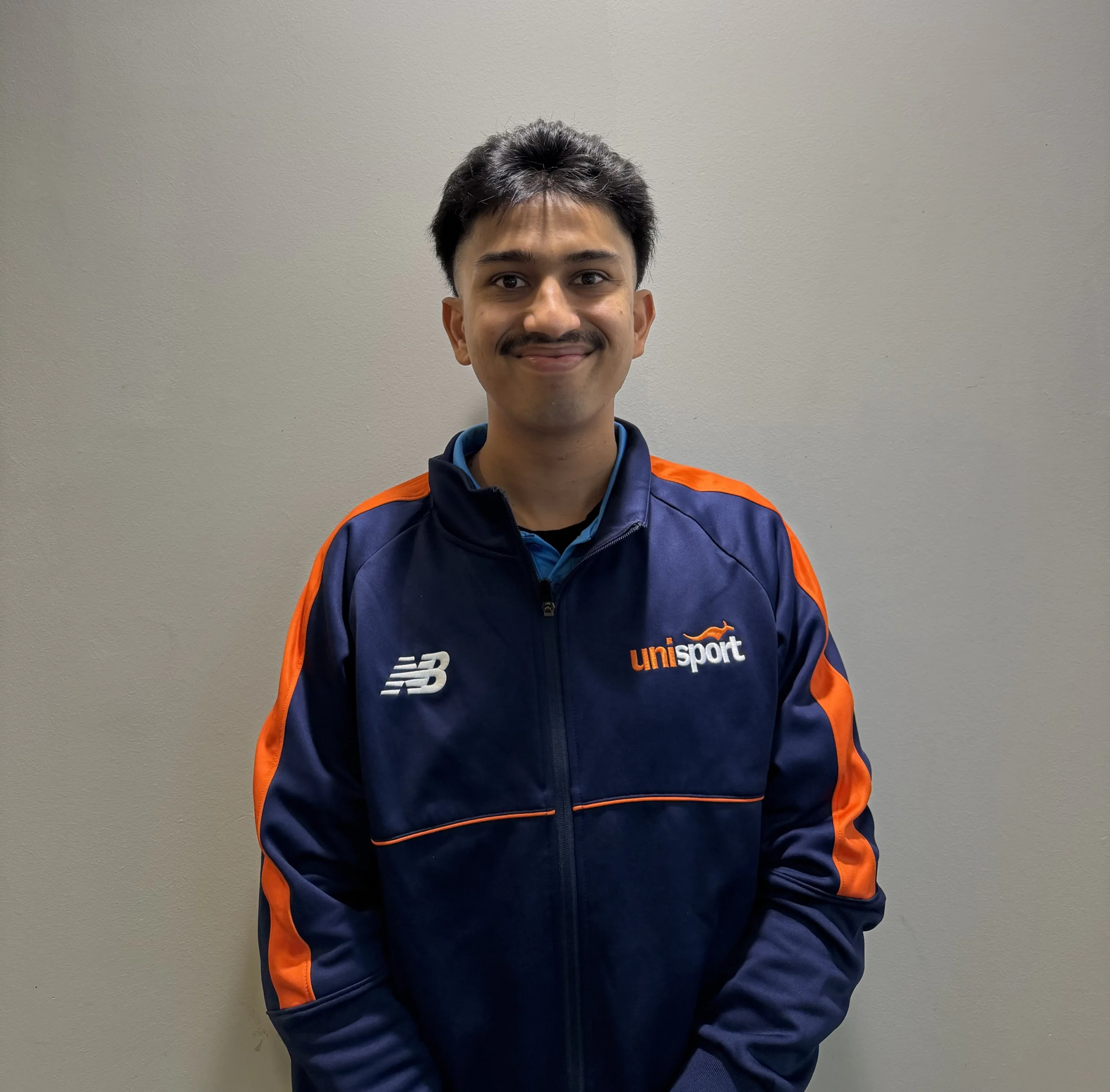 Front-facing man with short dark hair and a mustache, wearing a blue and orange sport jacket, standing against a gray wall.