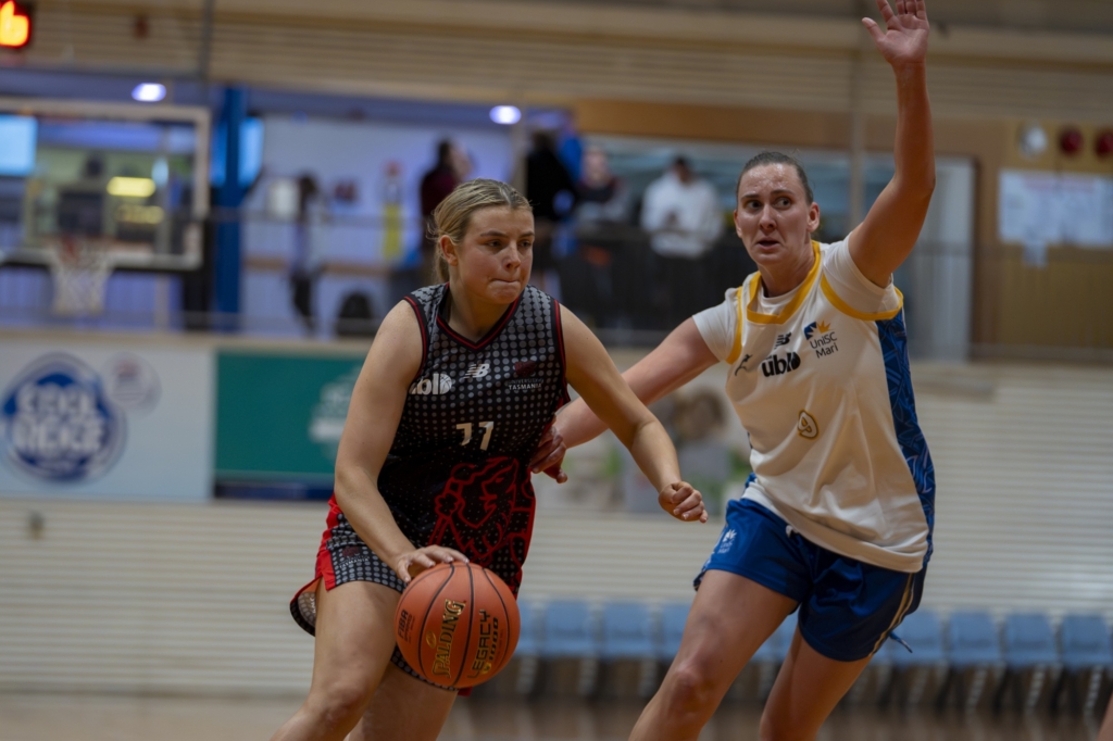 Two female basketball players compete for the ball on an indoor court, one dribbling in a black-red uniform while the other reaches in a white-blue jersey with arm raised for defense/shot block.