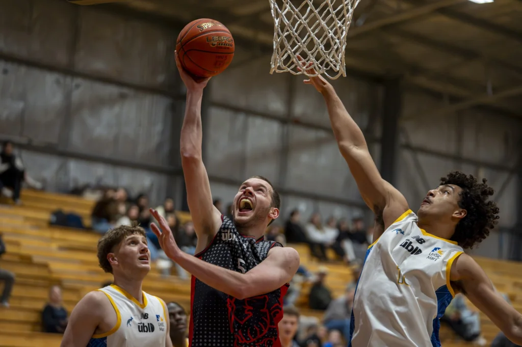 Basketball players jump near the hoop, one reaching for the ball as others guard; indoor gym with wooden bleachers in background.