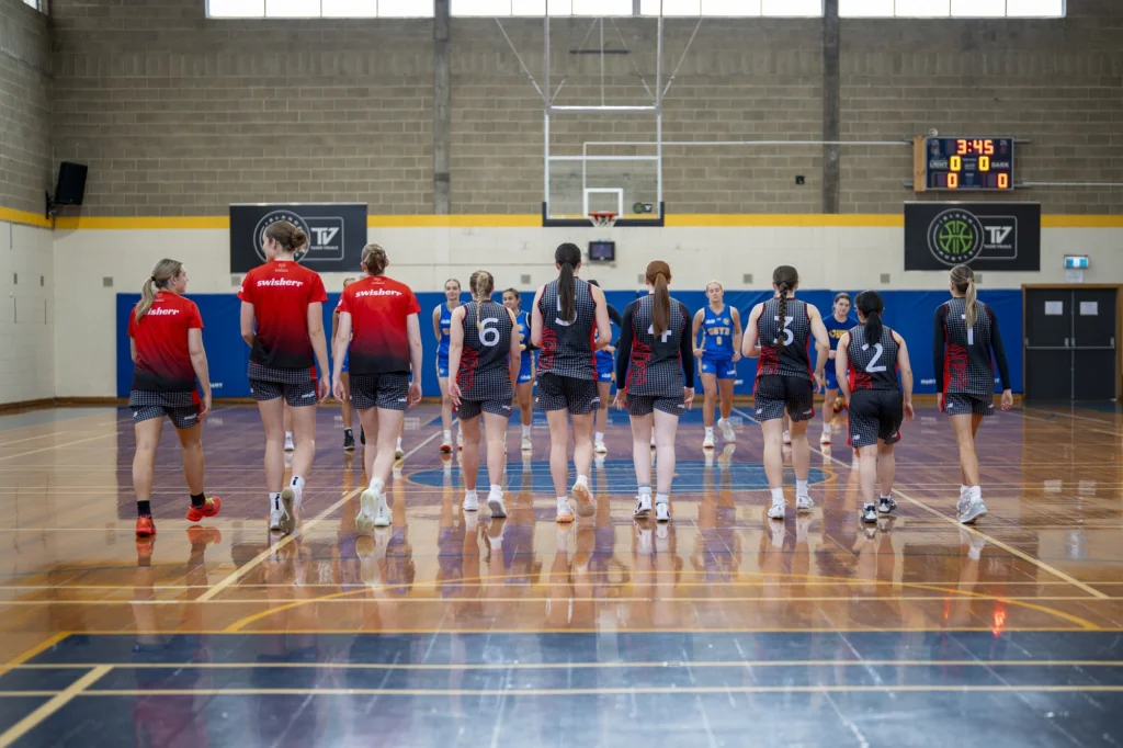 Female basketball players in red and blue uniforms walk toward the center of an indoor court, backs to camera.