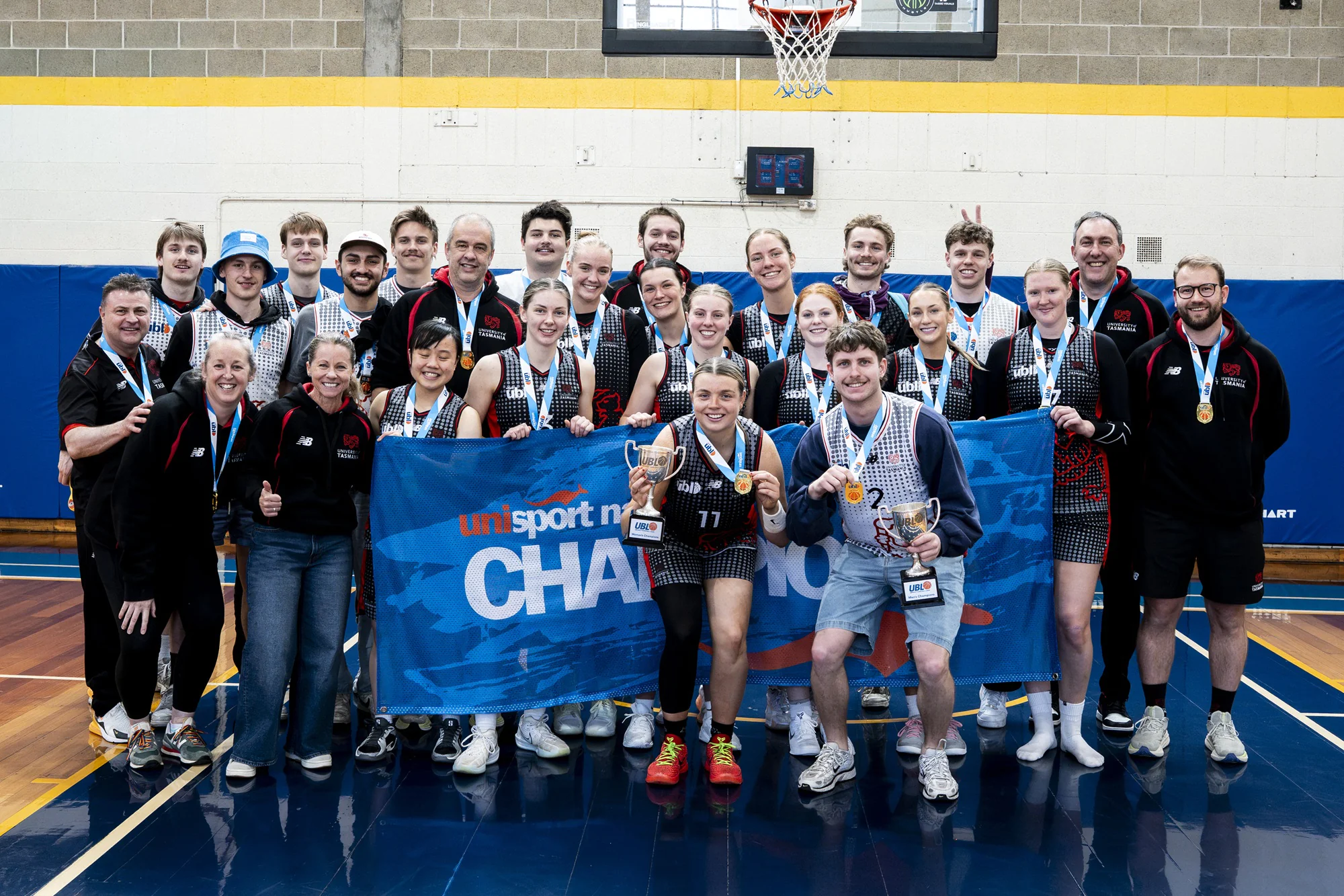 Group of athletes posing in a gym with medals and trophies after a competition, banner behind them reading 'UNI sport' and 'CHAMPIONS'