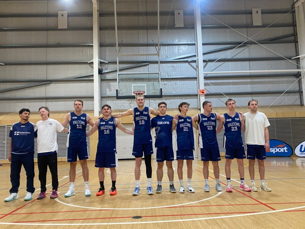 A line of male basketball players in blue Falcons jerseys pose arm-in-arm with coaches in a gymnasium.