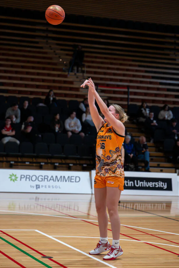 Female basketball player in an orange uniform taking a jump shot on an indoor court, with spectators in the stands behind her.