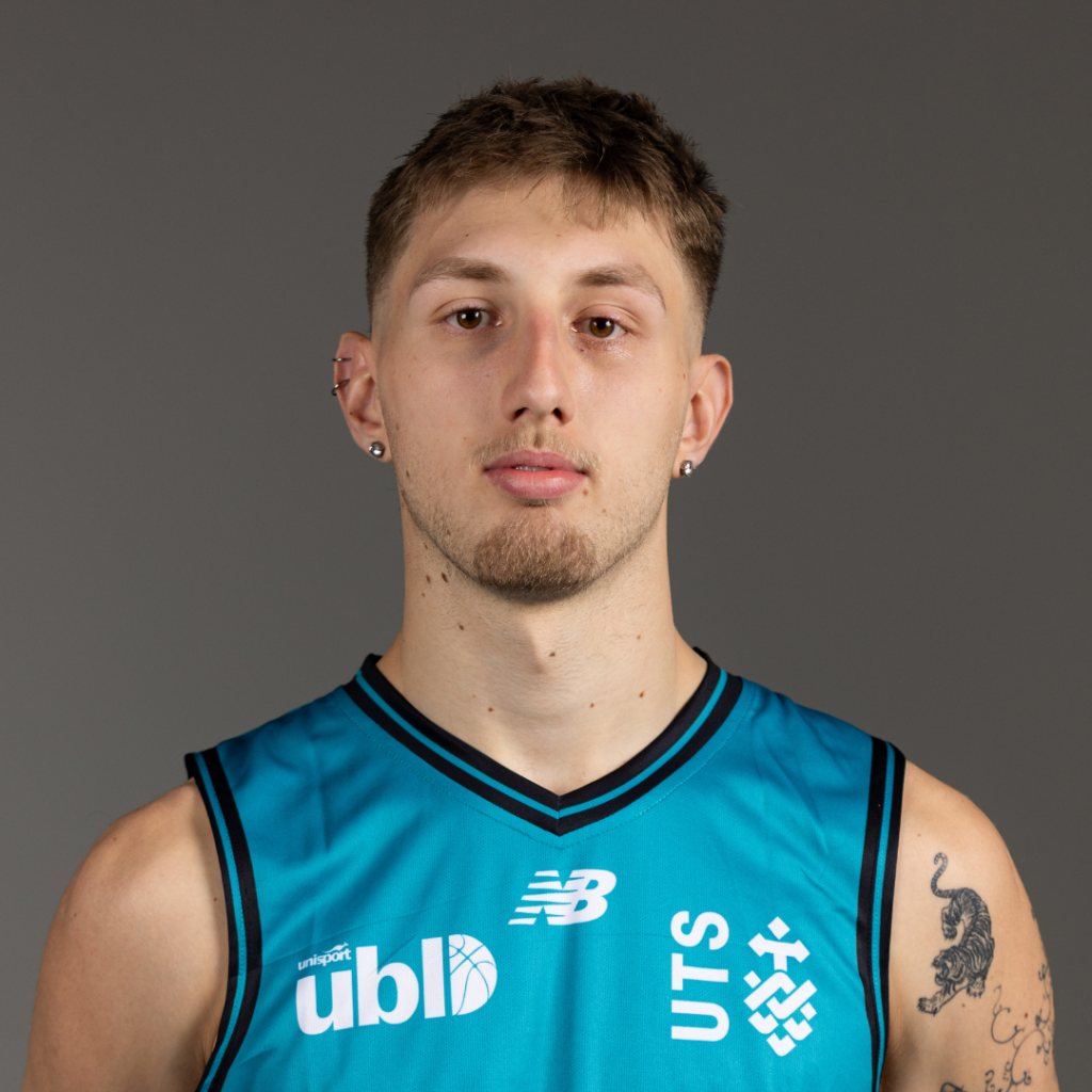 Portrait of a young man in a turquoise basketball jersey facing the camera with a neutral expression, tattoos visible on one arm.