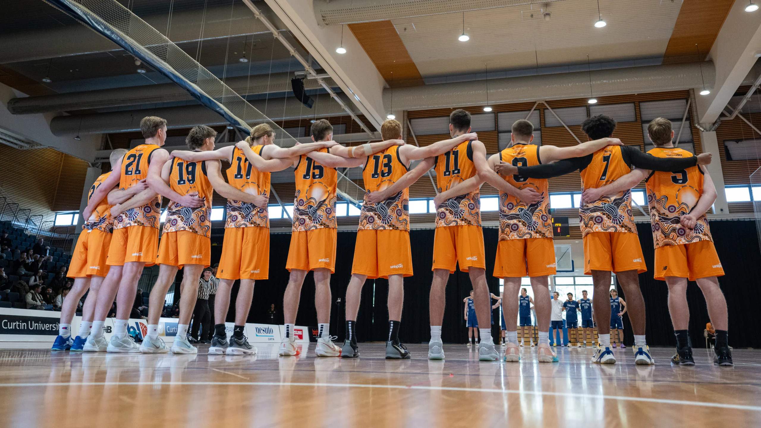 Basketball players in orange jerseys form a team huddle with arms around each other in a gymnasium.