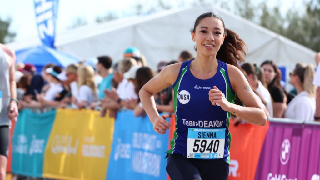 Female runner wearing a blue and green racing kit with bib 5940 (Sienna), sprinting along a barrier-lined course as cheering spectators watch behind.