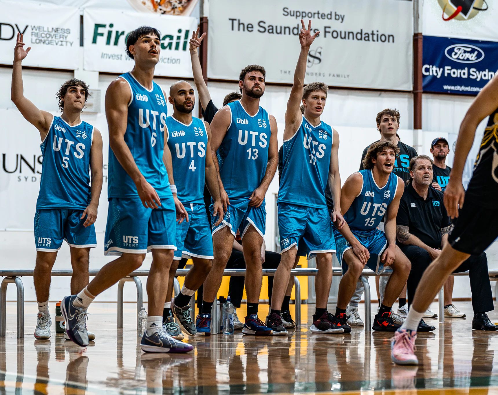 Basketball players in blue UTS uniforms stand along the bench as they raise their hands during a game in an indoor court with banners behind them.
