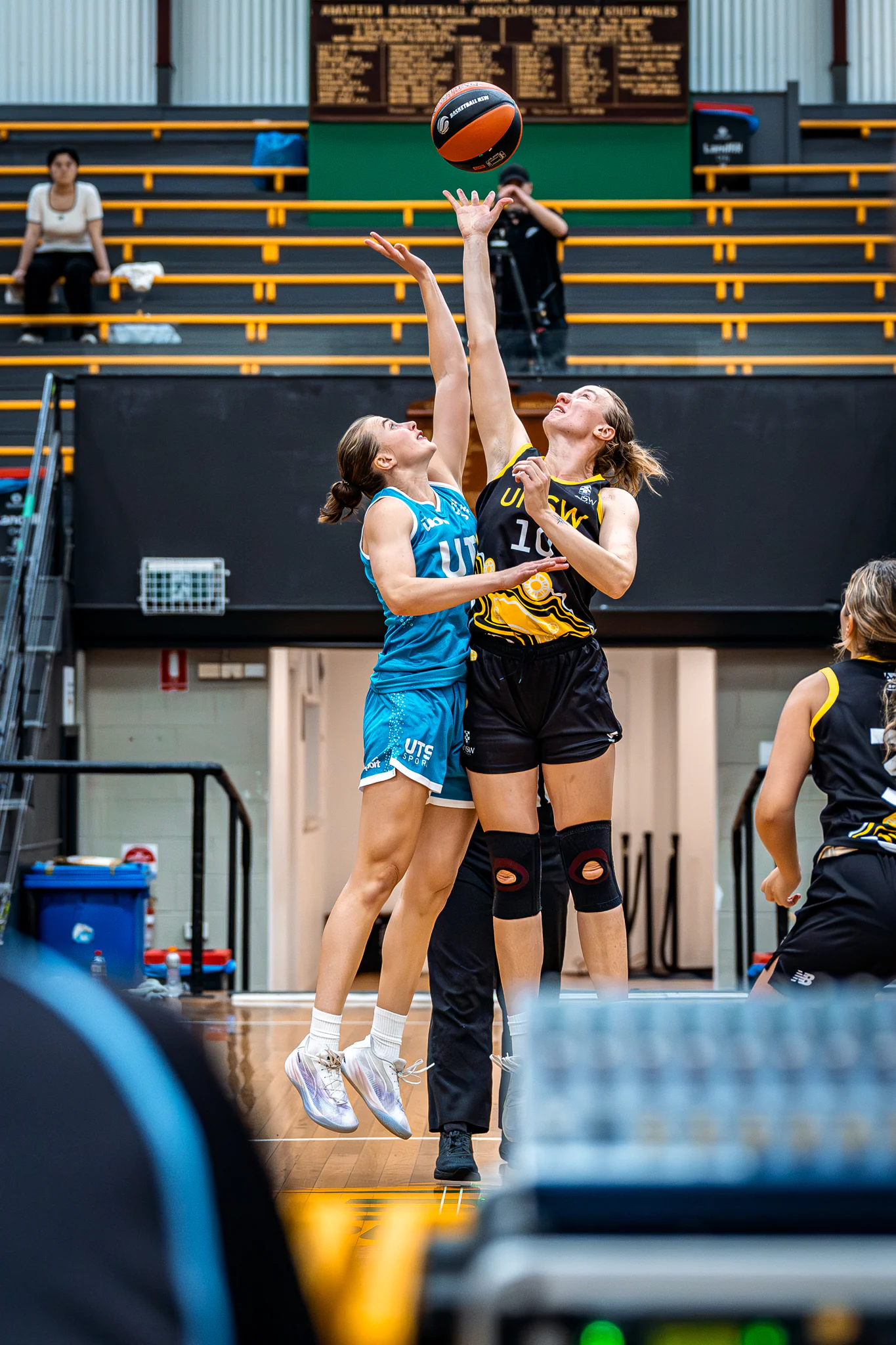 Two female basketball players jump to reach a ball during opening tip-off in a gym, one in blue and one in black-yellow uniforms.
