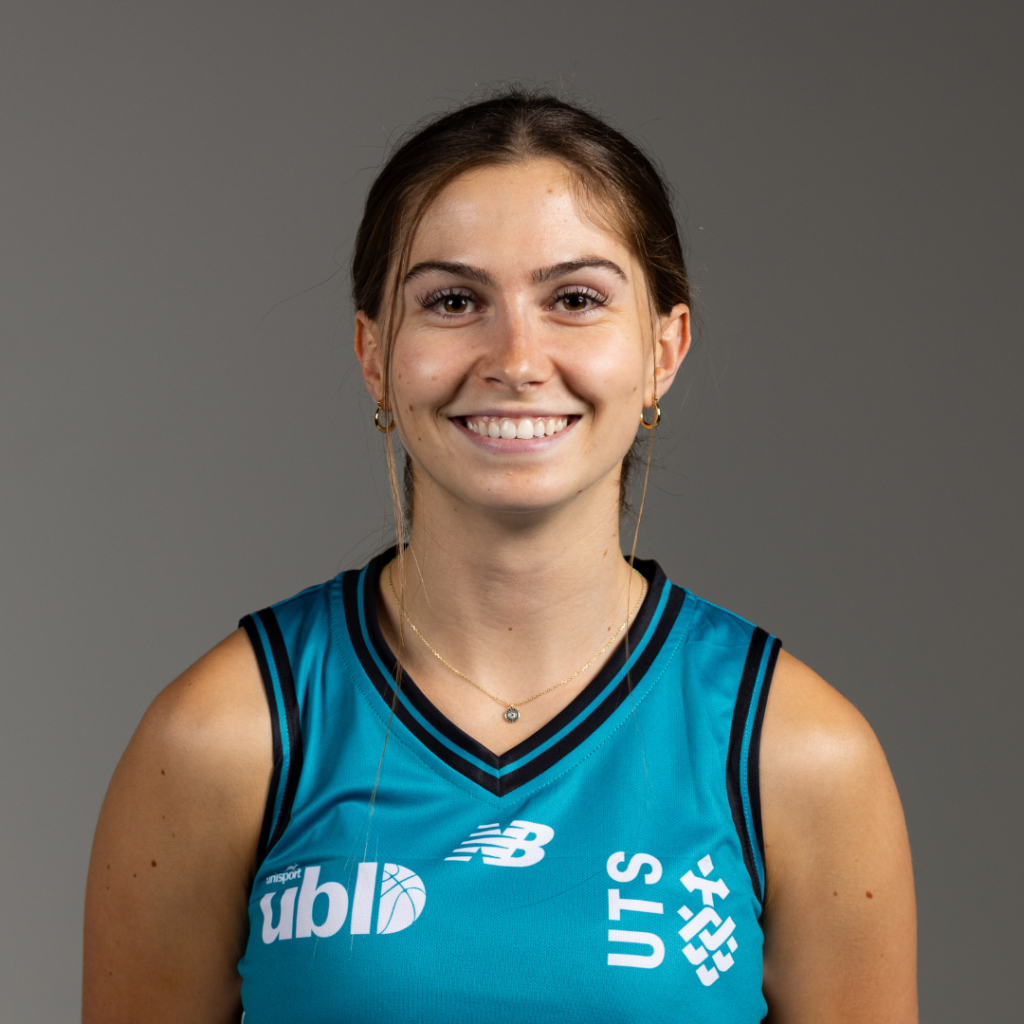 Smiling woman in teal athletic jersey with logos, facing camera against a neutral gray backdrop, wearing hoop earrings and a delicate necklace.