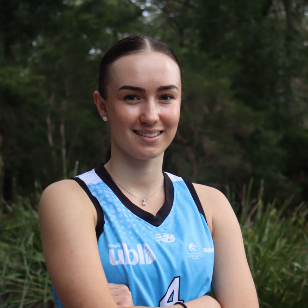 Smiling young woman in a blue athletic jersey with arms crossed, outdoors in a leafy park setting.