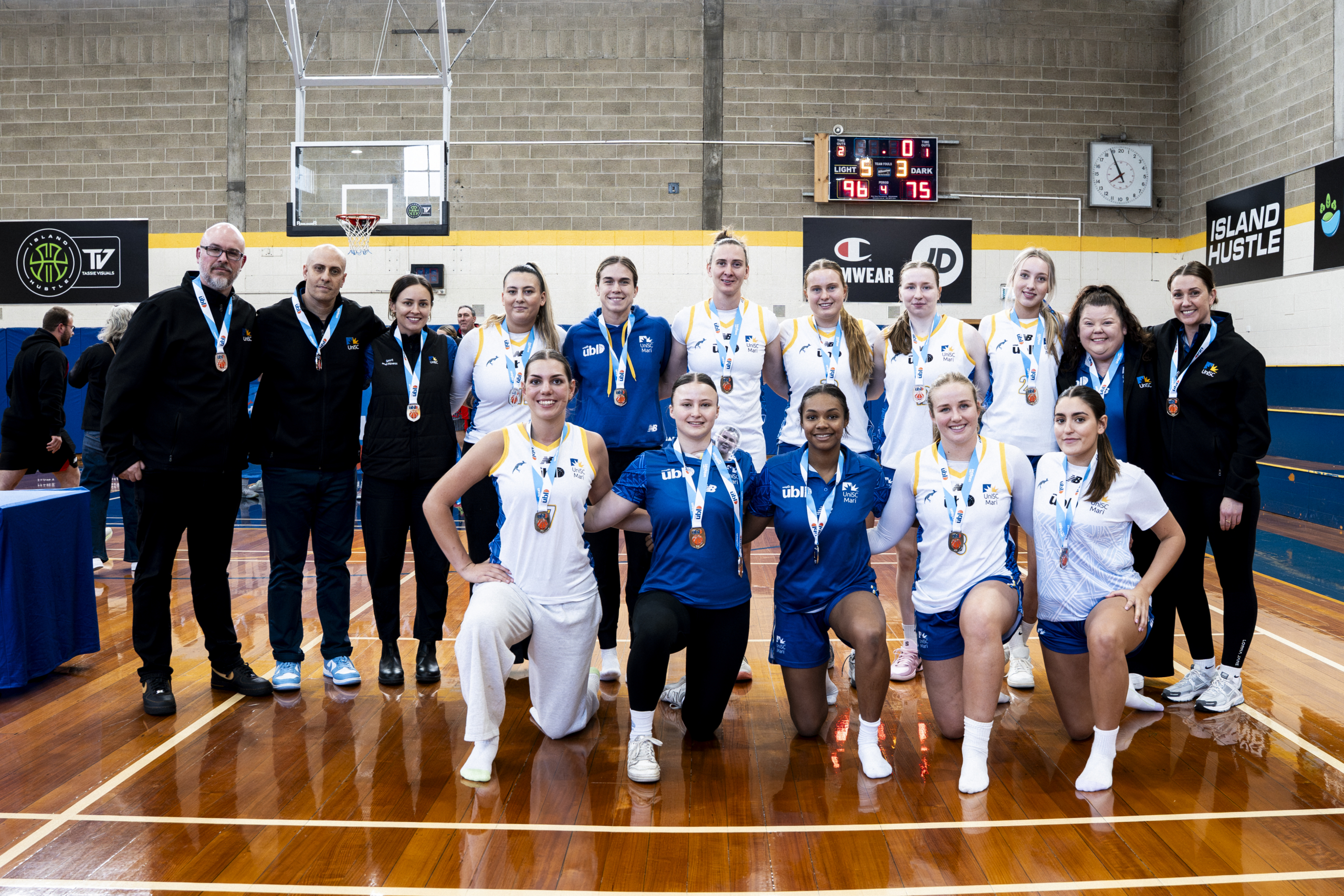 Group of basketball players in blue and white uniforms wearing medals, posing with coaches in a gymnasium.