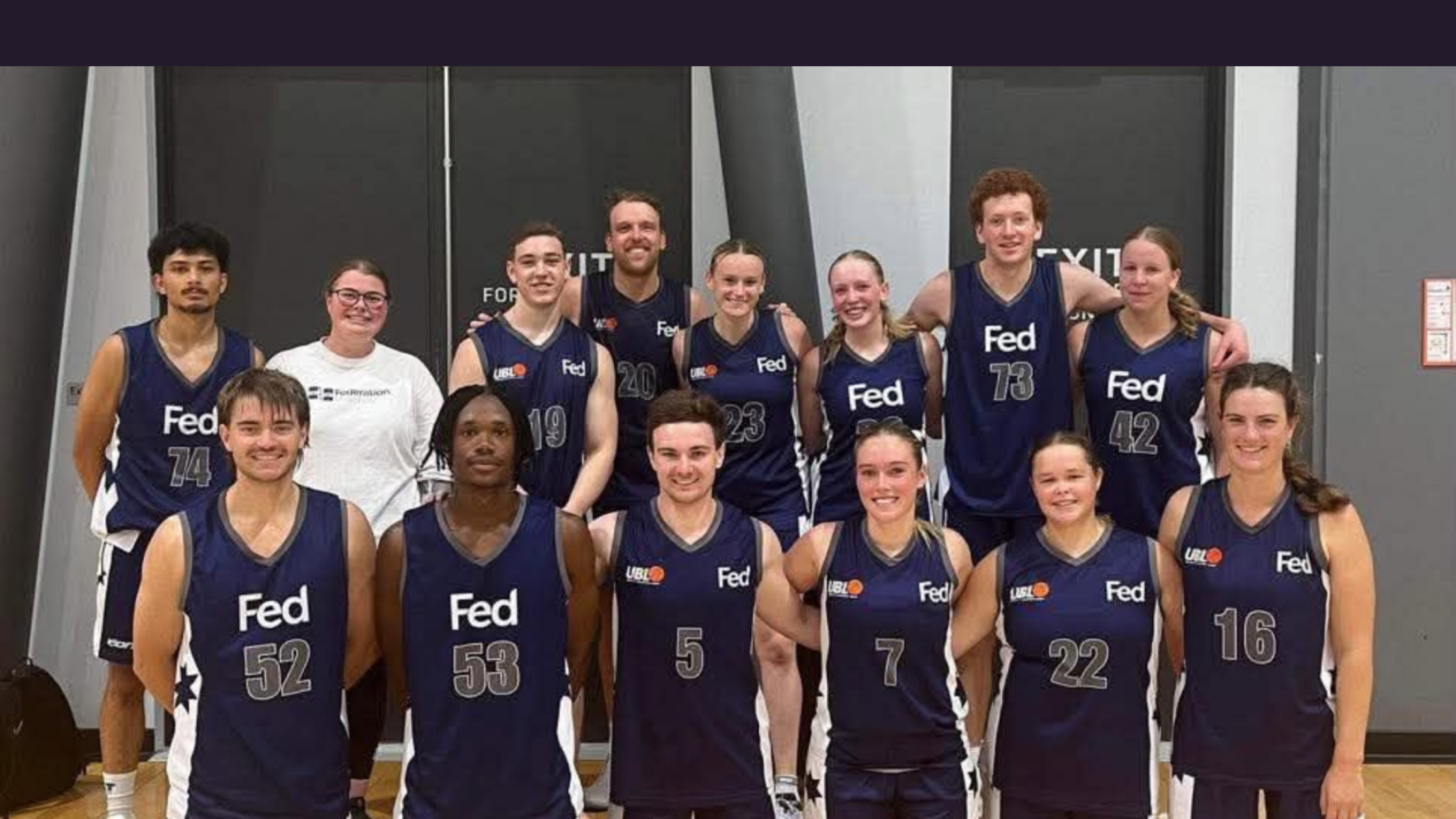 Group of basketball players in navy uniforms with the 'Fed' logo posing together in a gym, many smiling at the camera.
