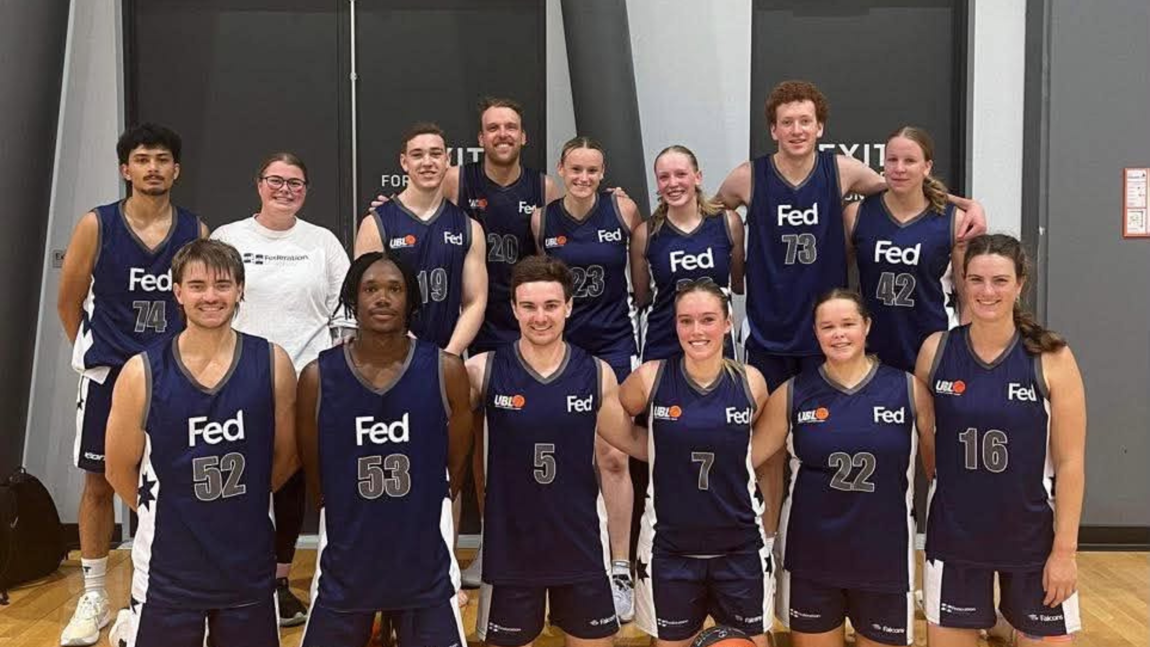 Group of basketball players in navy uniforms posing for a team photo in a gym, smiling at the camera.
