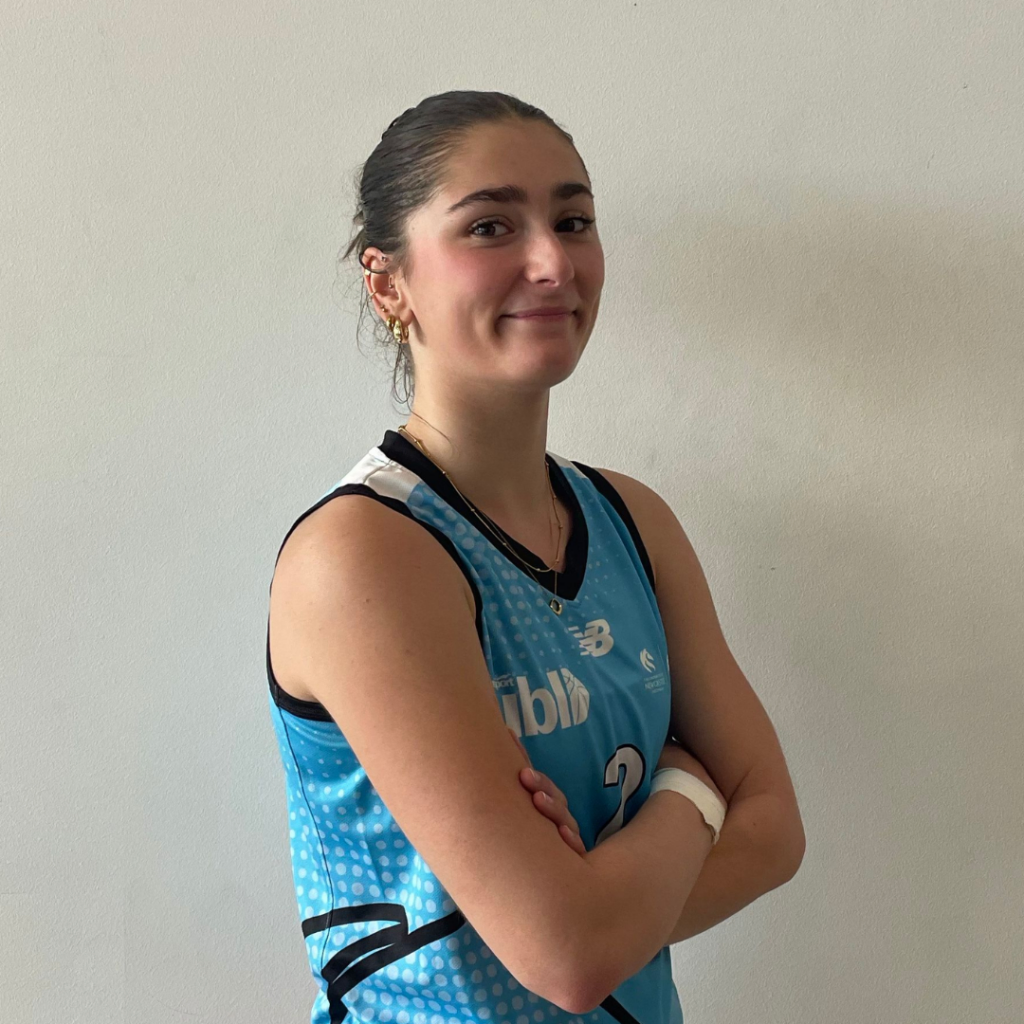 Athlete in a blue sleeveless jersey with arms crossed, smiling at the camera against a plain wall.