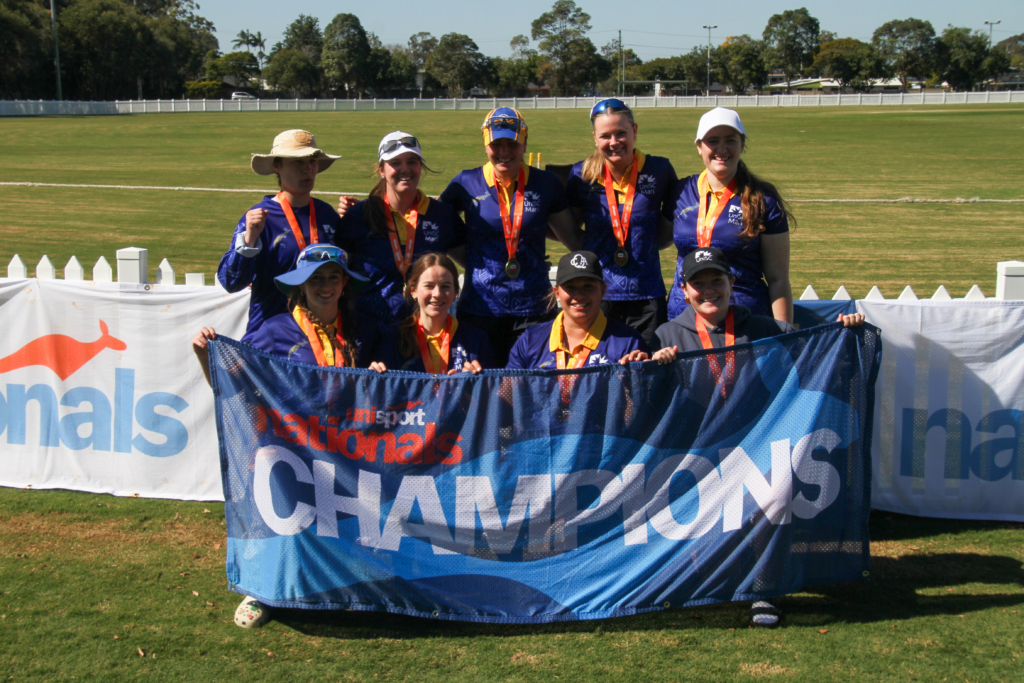A group of female athletes in blue uniforms posing with medals around their necks and a large blue banner reading 'NATION... CHAMPIONS' on the field behind them.