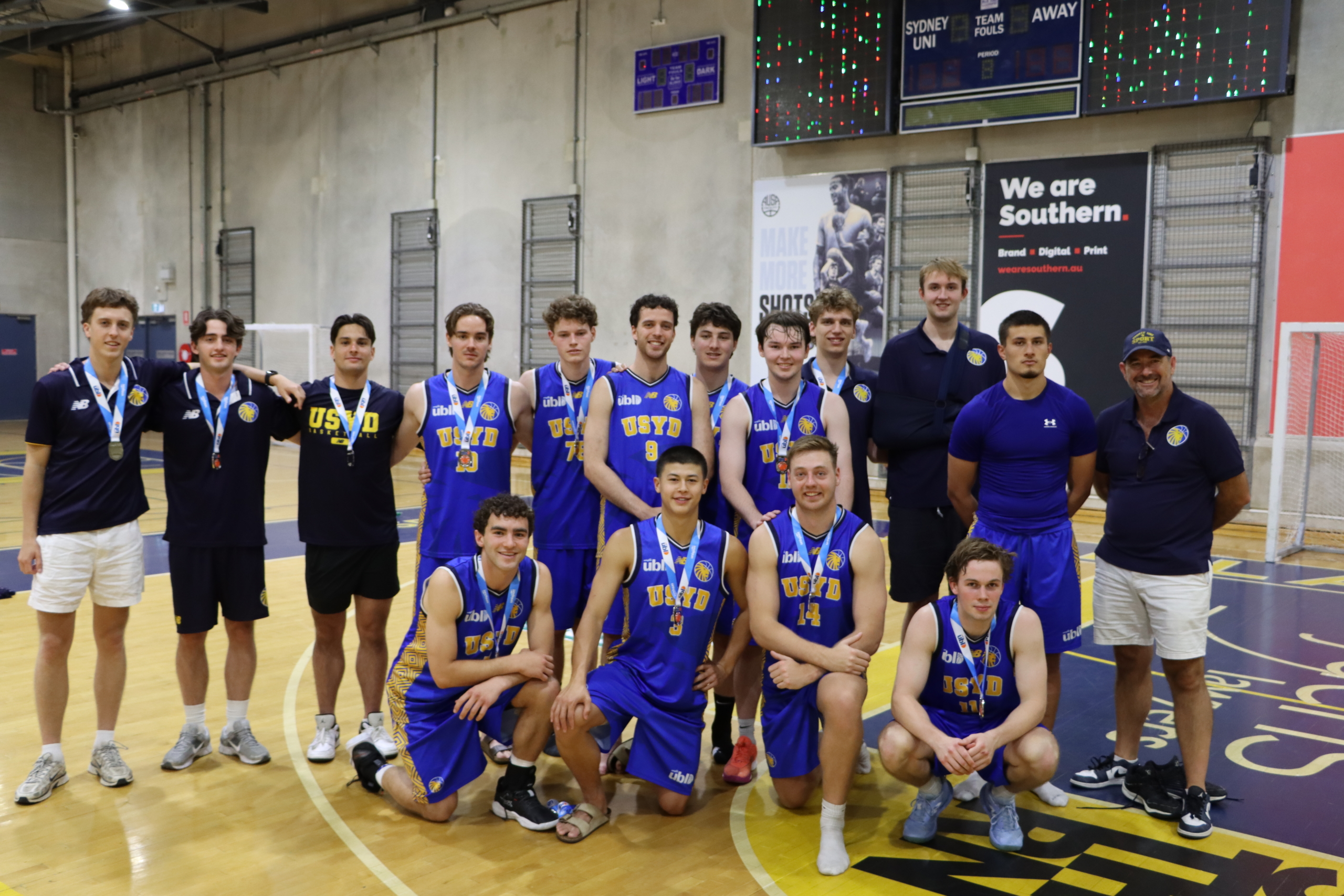 Group of young basketball players in blue uniforms posing for a team photo in a gym, with medals around their necks.