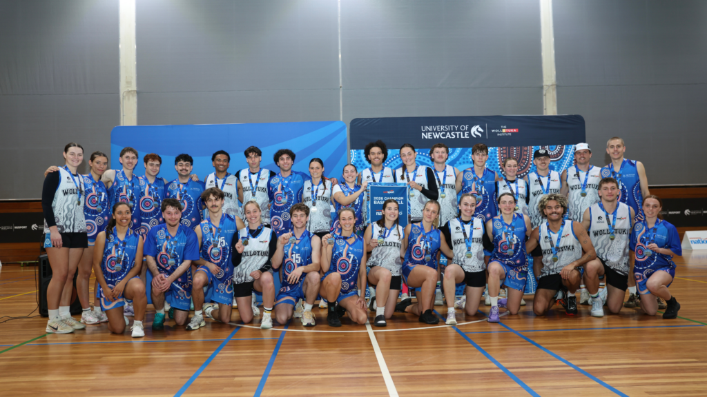 Group of athletes in blue and white patterned uniforms posing as a team on a gym floor, medals around their necks after a game or tournament, with banners in the background.