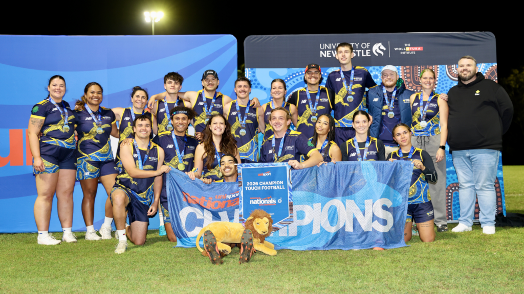 A large group of athletes in matching blue and yellow uniforms posing with medals on a blue backdrop at night, holding banners.