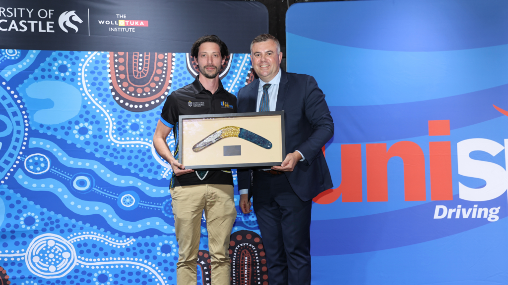 Two men pose with a framed beaded boomerang award at a university event, against a blue patterned backdrop.
