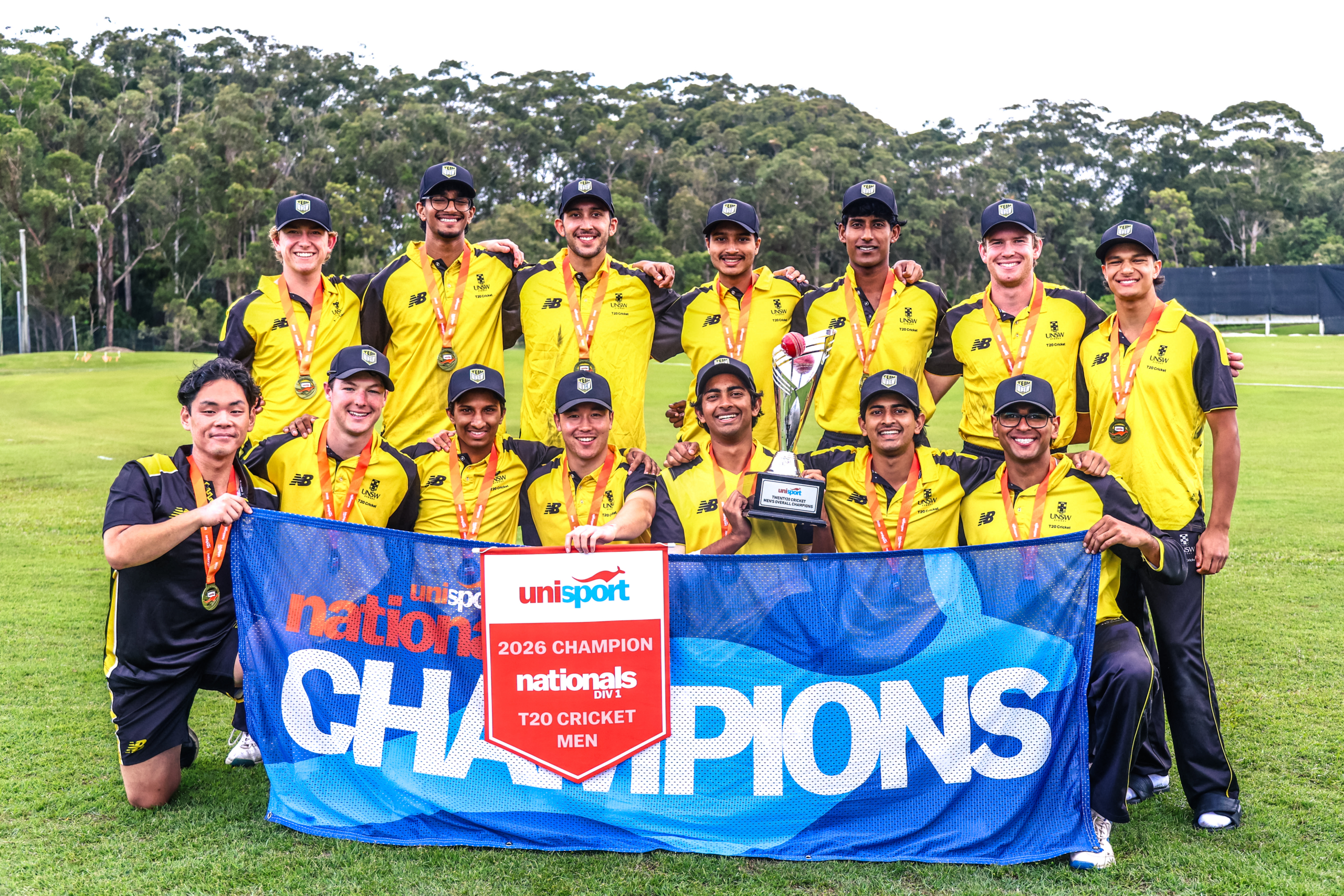 Cricket team in yellow uniforms with medals posing on a field, holding a blue banner that reads NATIONALS CHAMPIONS with a red champion badge visible below.