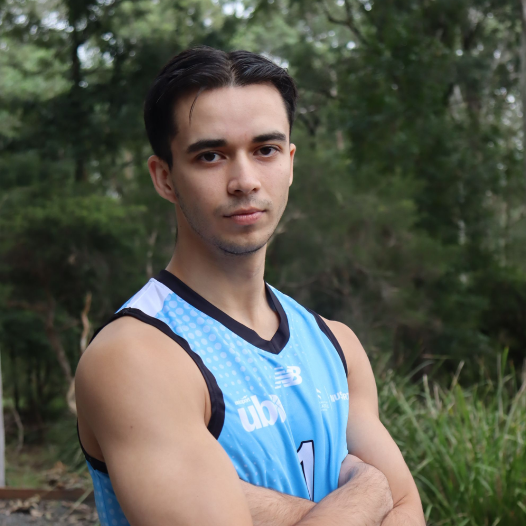 Young man with short dark hair posing outdoors with arms crossed in a blue athletic jersey.