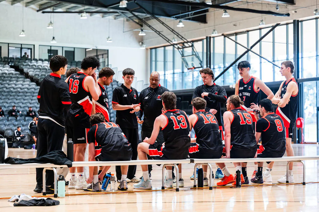 Basketball players in black uniforms sit on a bench during a timeout, listening to their coach on a gym floor with bleachers in the background.