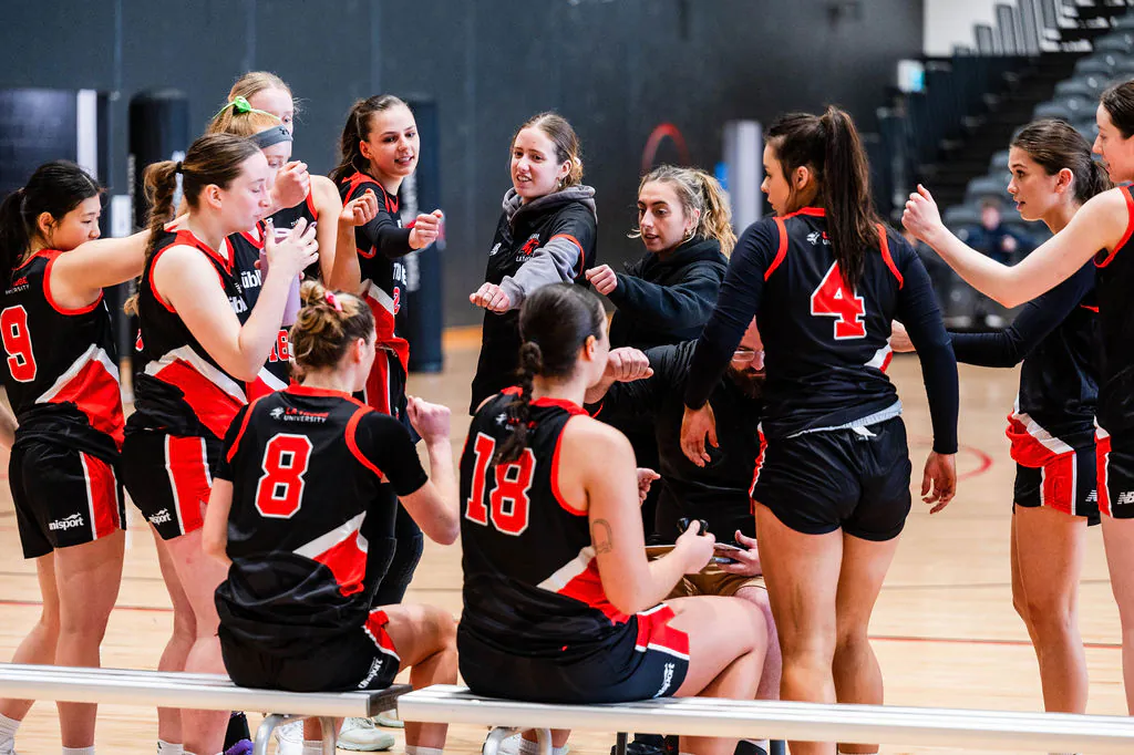 Girls’ sports team huddle on an indoor court, red-and-black uniforms, high-fiving and cheering together.