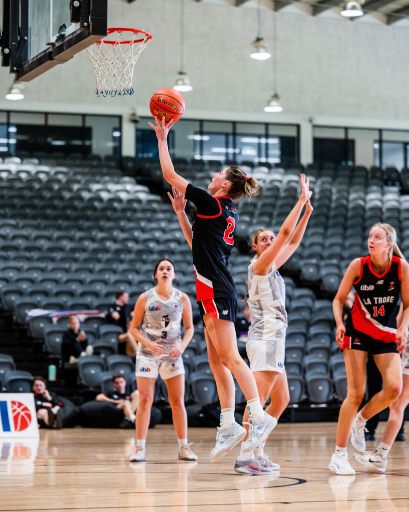 Female basketball player in black jersey (#2) jumps for a layup toward the hoop as defenders reach up, indoors on a polished court with empty stands behind.