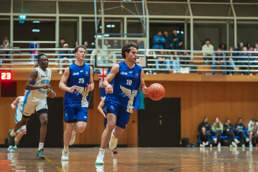 Basketball game: two players in blue uniforms dribble toward the basket while spectators watch from stands behind a railing.