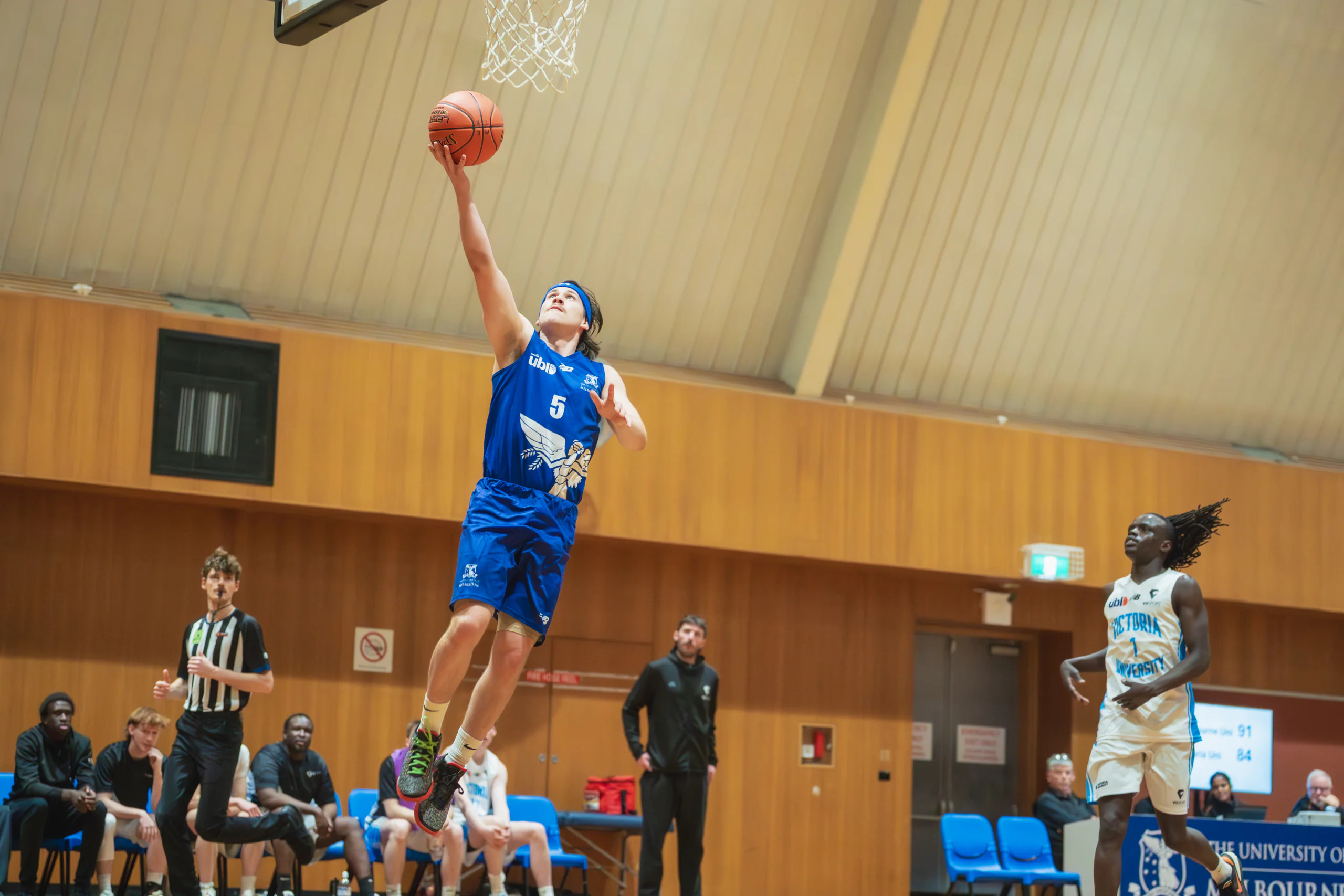 Basketball player in a blue uniform leaps toward the hoop for a shot in an indoor gym, with teammates and spectators watching.