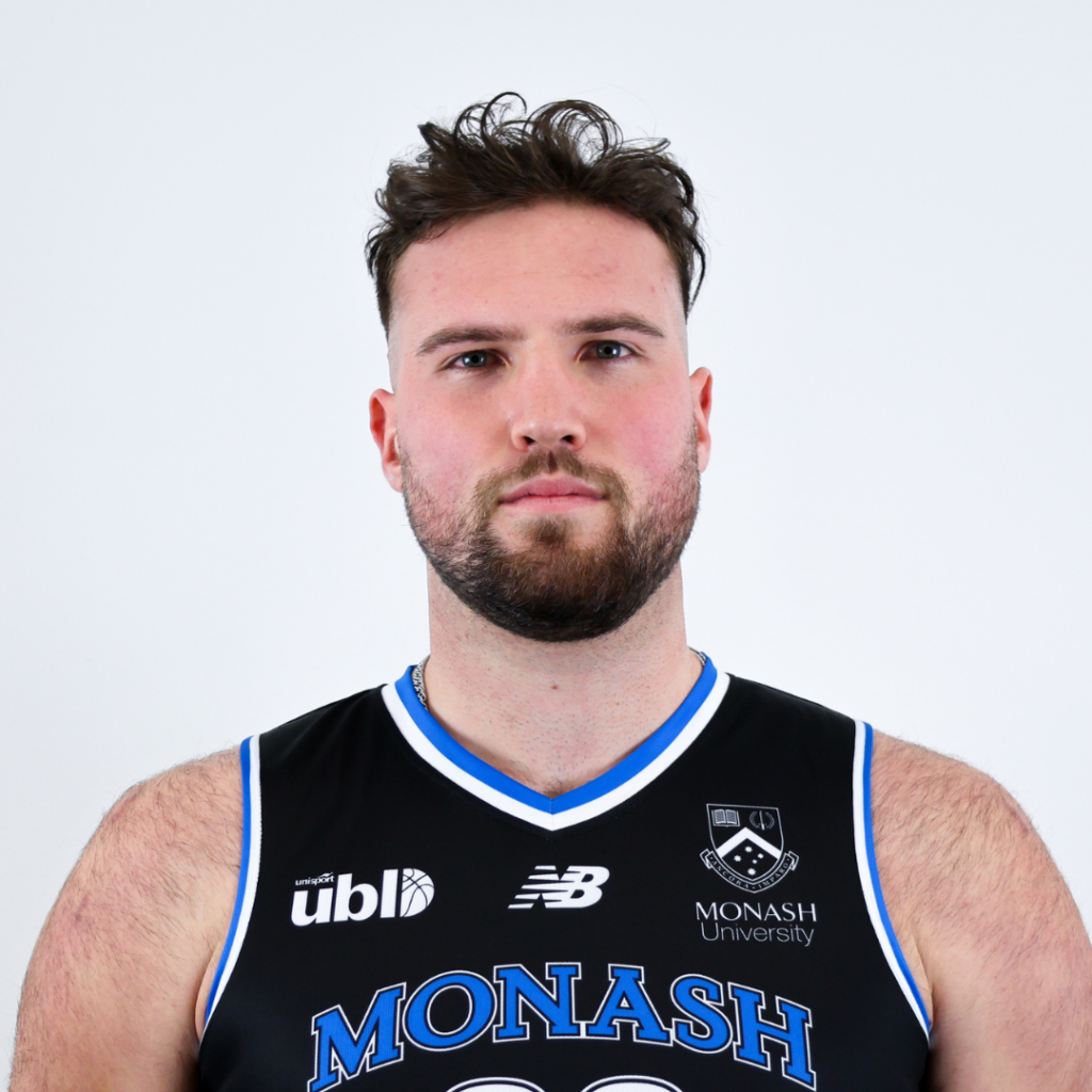 Portrait of a man with short dark hair and a beard, wearing a black Monash University basketball jersey and looking at the camera.