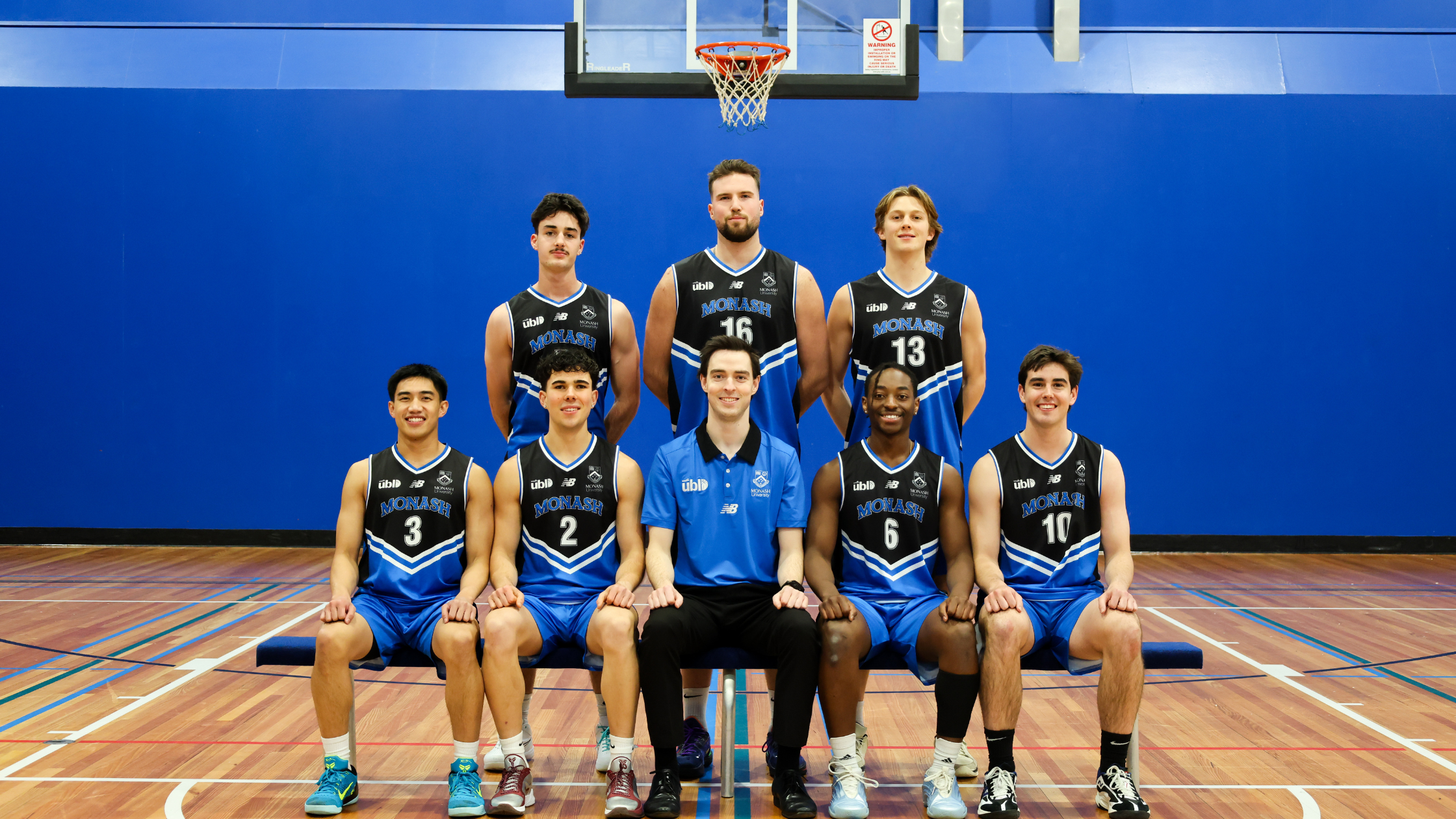 Group photo of Monash basketball team posing in gym; six players kneeling in front, three standing behind, blue wall background.
