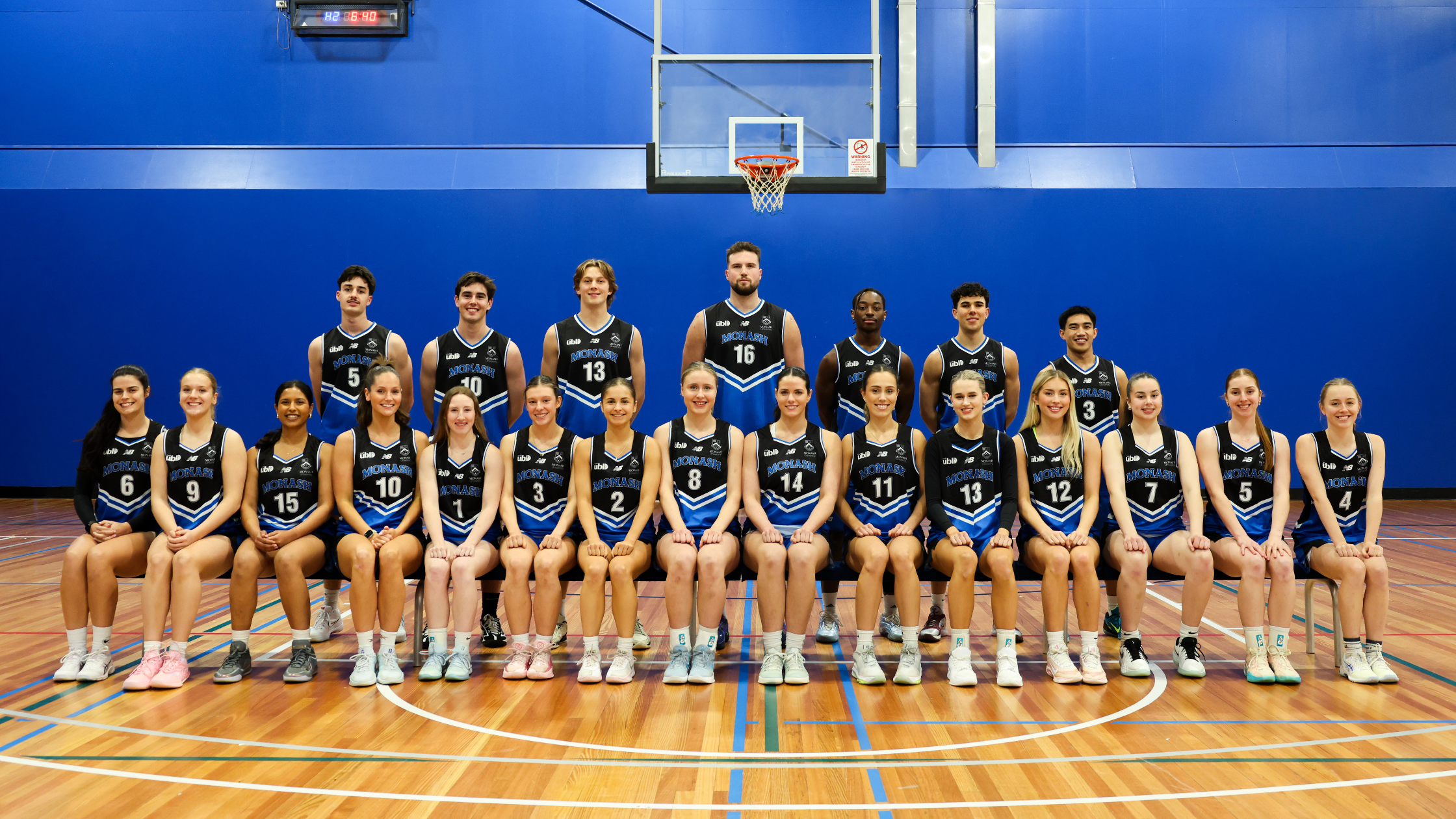 Group photo of a basketball team in blue and black uniforms posing in a gym, with players seated in front row and standing behind on the court.